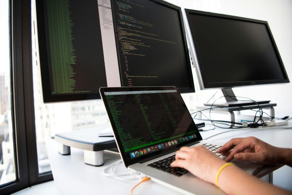 Haende einer Person tippen an einem Laptop mit Code vor zwei Monitoren