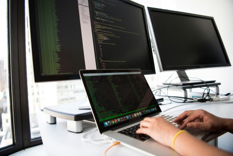 Hände einer Person tippen an einem Laptop mit Code vor zwei Monitoren
